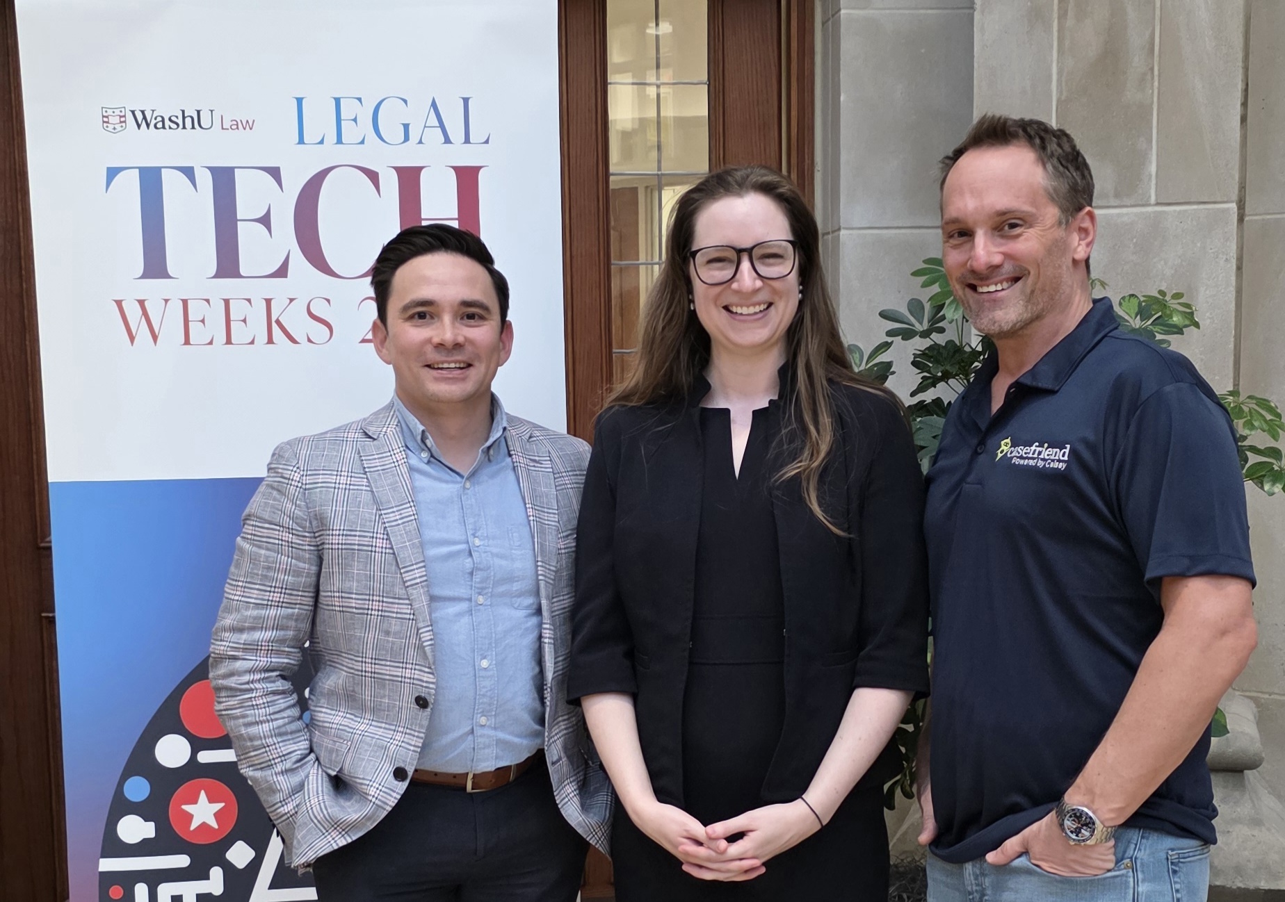 Three individuals standing together in front of a WashU Law ‘Legal Tech Weeks’ event banner, posing for a photo inside a building with stone walls and potted plants.